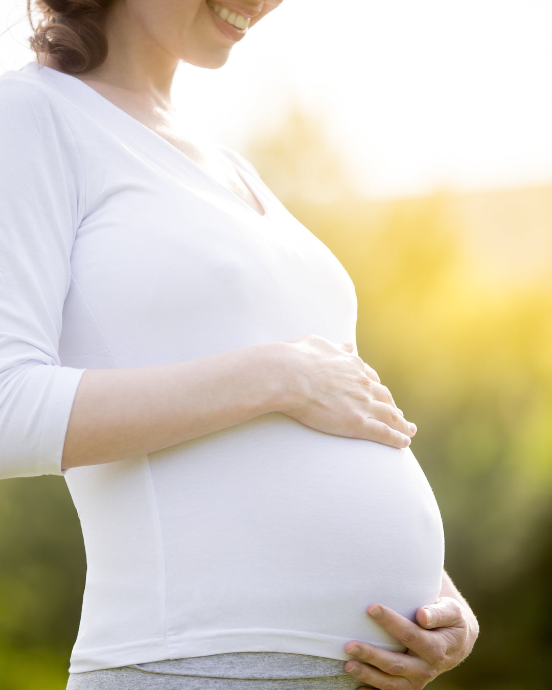 Pregnant woman wearing a white long-sleeve V-neck top and gray leggings, cradling her baby bump outdoors with warm sunlight and a blurred green background.