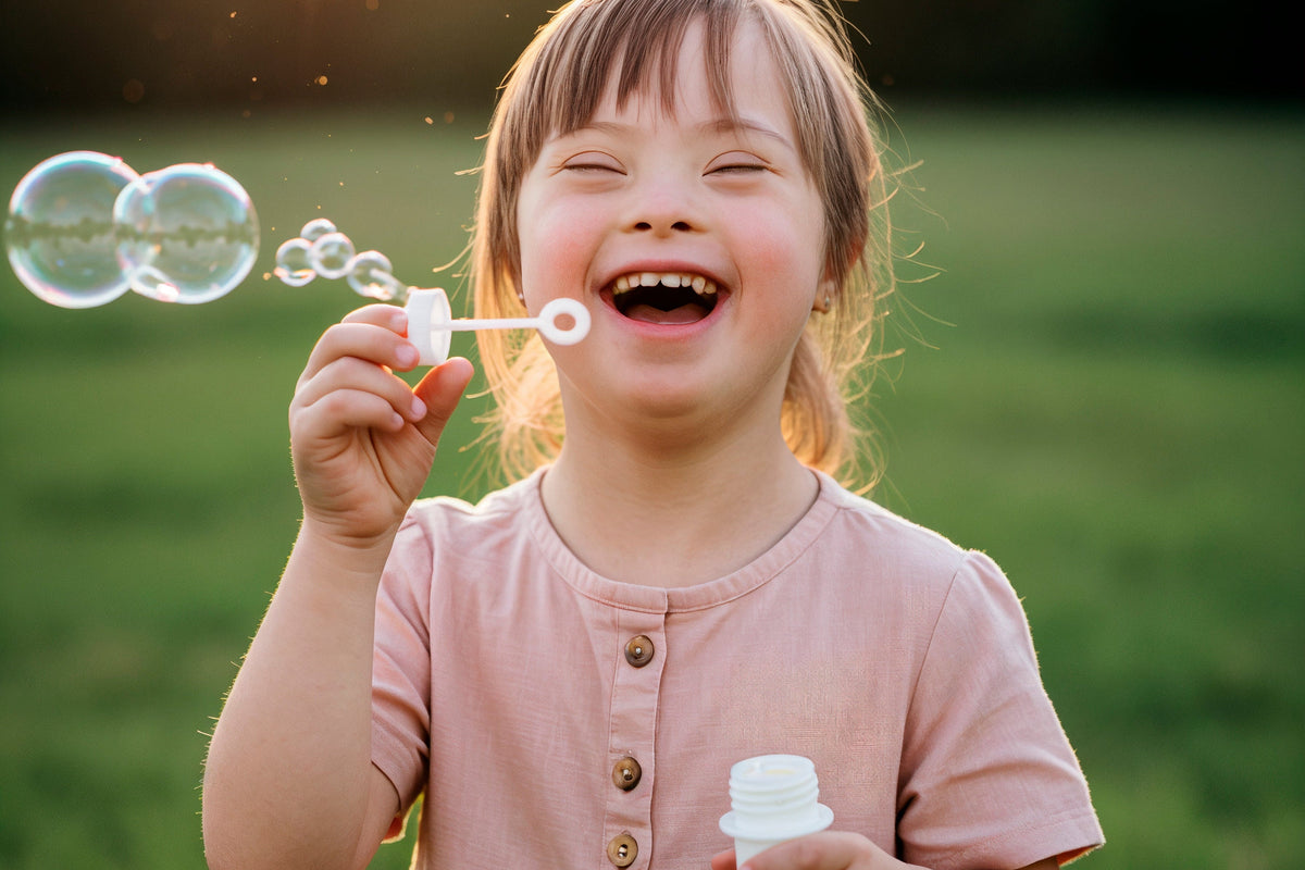 A young girl with Down syndrome is smiling and laughing while blowing bubbles in an outdoor grassy field during golden hour. She is wearing a dusty pink short-sleeve dress and holding a bubble bottle.