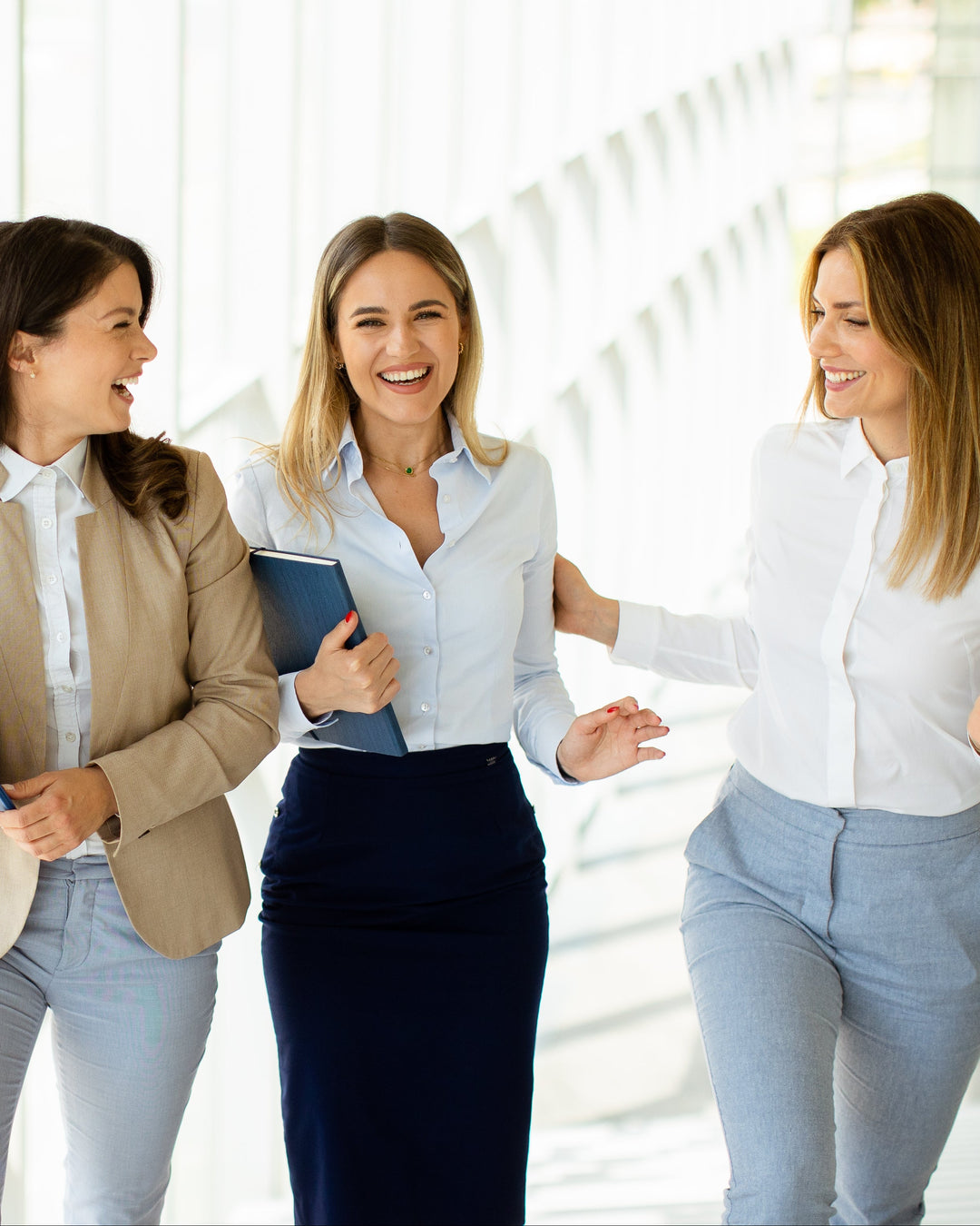 Three professional women walking and smiling in a bright, modern office corridor with large windows.